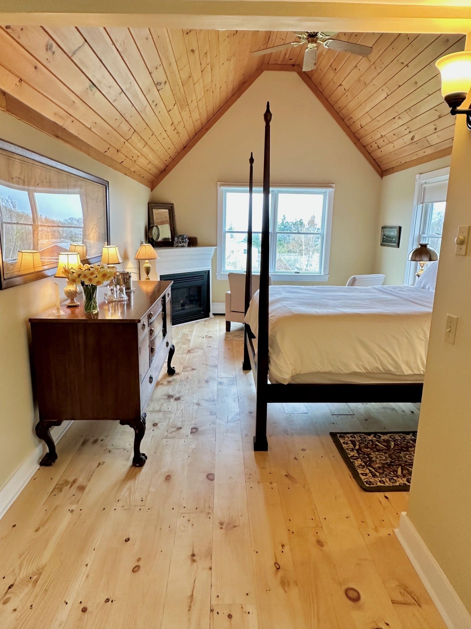 A view of the room from the entry showing beautiful wood floors and high ceiling, colors are warm and golden. A high posted bed faces a wall with a framed nautical chart, a corner fireplace near the back wall, with windows in the back and the side of the room.