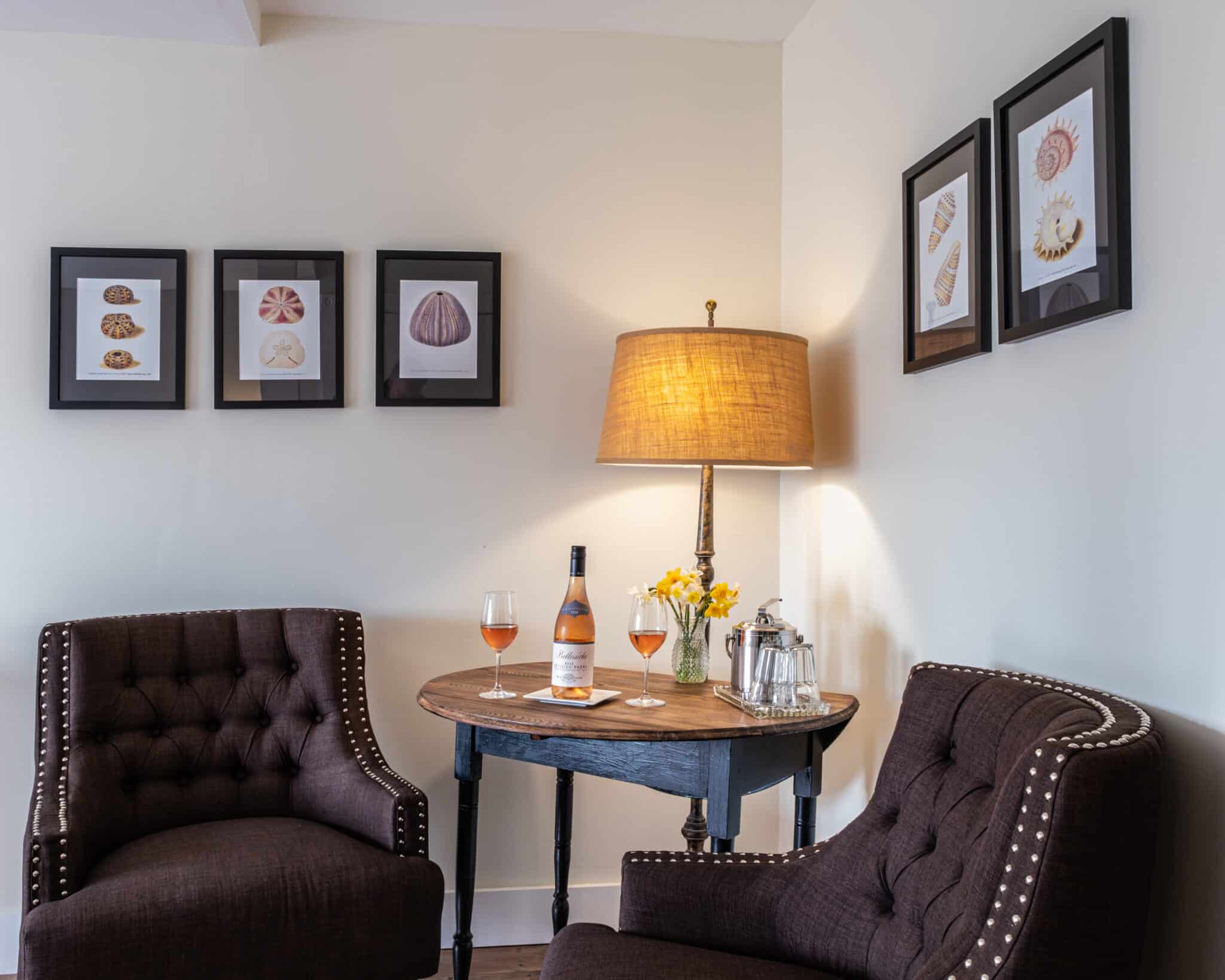 two brown chairs, a table with lamp on, bottle of wine and glasses with shell prints on the wall