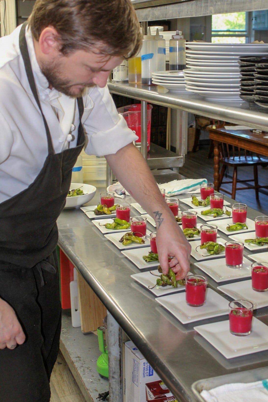 Chef Ross Moskwa plating numerous dishes for an event. Bright pink beet soup in glasses with vibrant green shishito pepper on the side