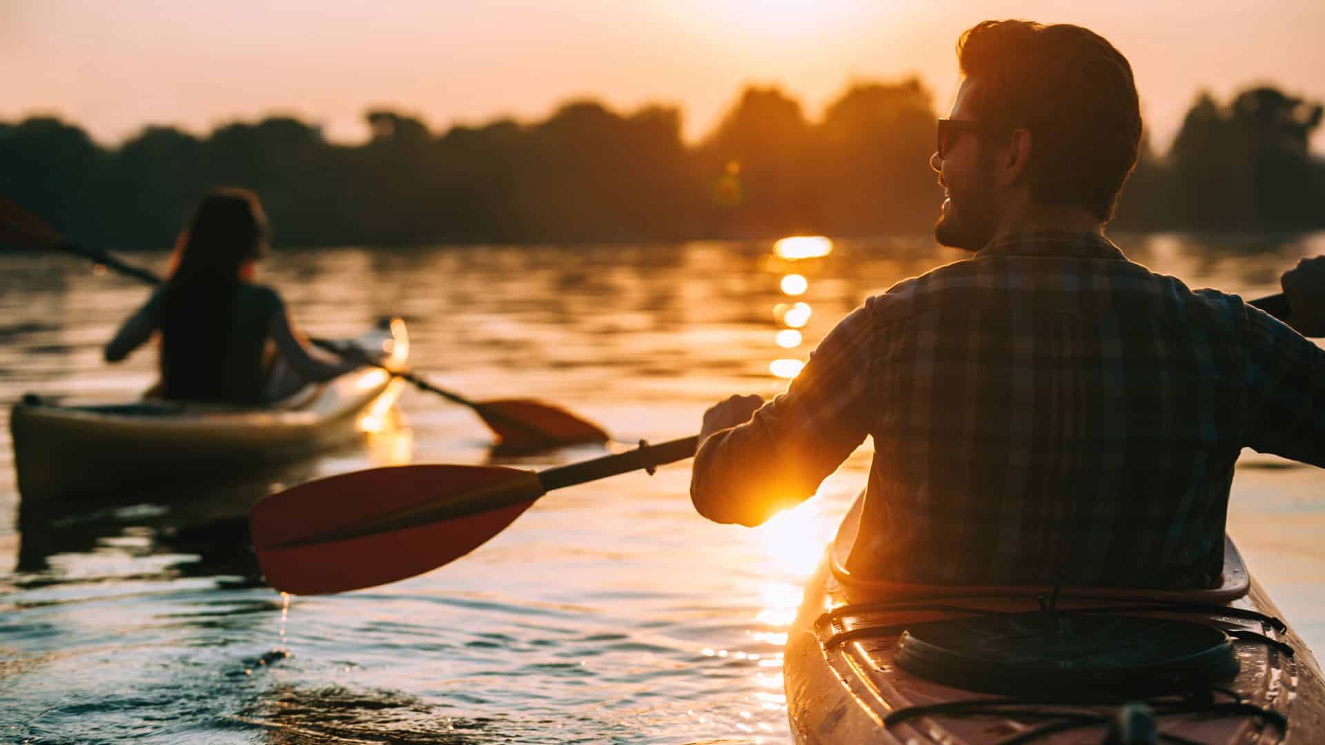 Two people kayaking on still water with trees and the sun in the background