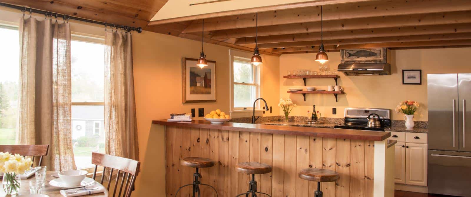 Large room with cream walls, wooden paneling, kitchen, stainless steel appliances, breakfast bar, three wood and iron stools, and dining area with wooden table and chairs