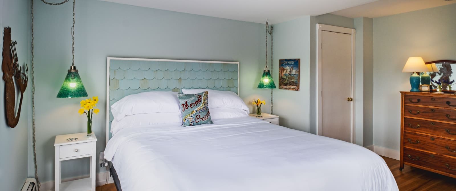 Large bedroom suite with light blue walls, hardwood flooring, white bedding, and green glass lamps hanging from the ceiling