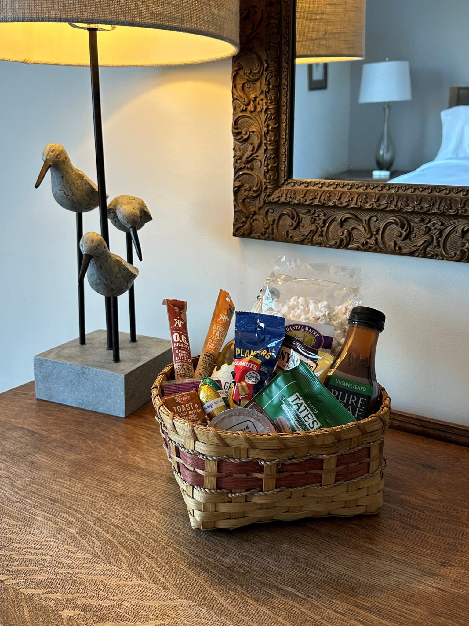 Basket filled with snacks and drinks set on a dresser with a lamp base of three birds in view and a mirror in which the bed and nightstand and lamp are reflected