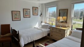 Twins beds with crisp white linens and wooden antique bed frames. Walls are off white with framed vintage bathing beauty Life Magazine covers. Wall lamps, dresser between bed, windows behind each bed. view of one orange leather chair at the end of the bed.