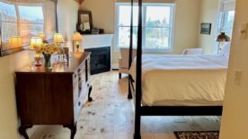 A view of the room from the entry showing beautiful wood floors and high ceiling, colors are warm and golden. A high posted bed faces a wall with a framed nautical chart, a corner fireplace near the back wall, with windows in the back and the side of the room.