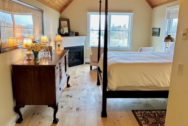 A view of the room from the entry showing beautiful wood floors and high ceiling, colors are warm and golden. A high posted bed faces a wall with a framed nautical chart, a corner fireplace near the back wall, with windows in the back and the side of the room.