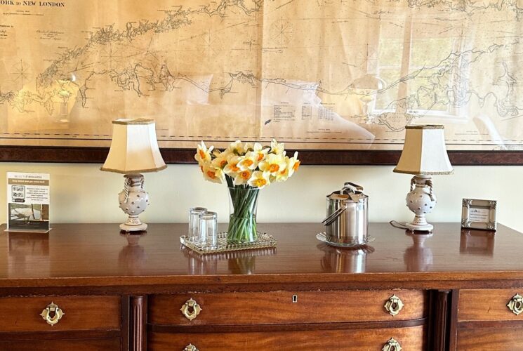 View of a large, beautiful antique dresser topped with daffodils, two small lamps, glassware and an ice bucket. A large framed, antique, nautical chart hangs on the wall.