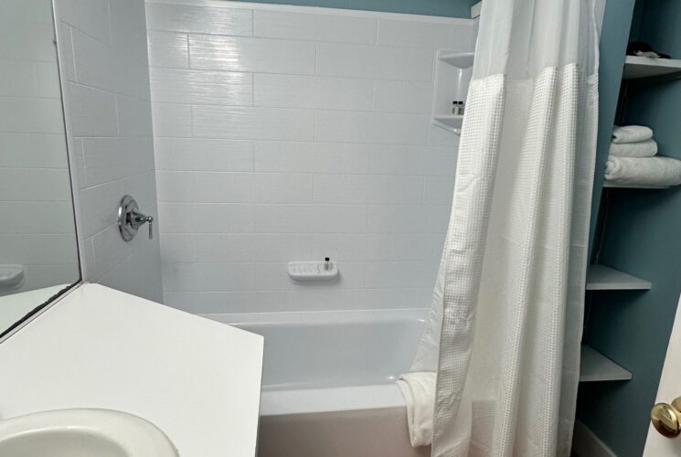 View into the bathroom. Blue walls, bright white shower tub unit, shelves to the side and wooden floors.