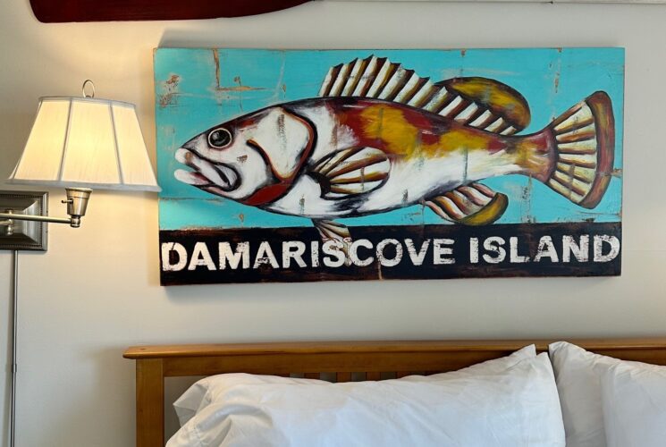 Close up view of the wall behind the bed with a large fish painting and the words Damariscove Island. A white and brown oar is above the painting and the king bed below.
