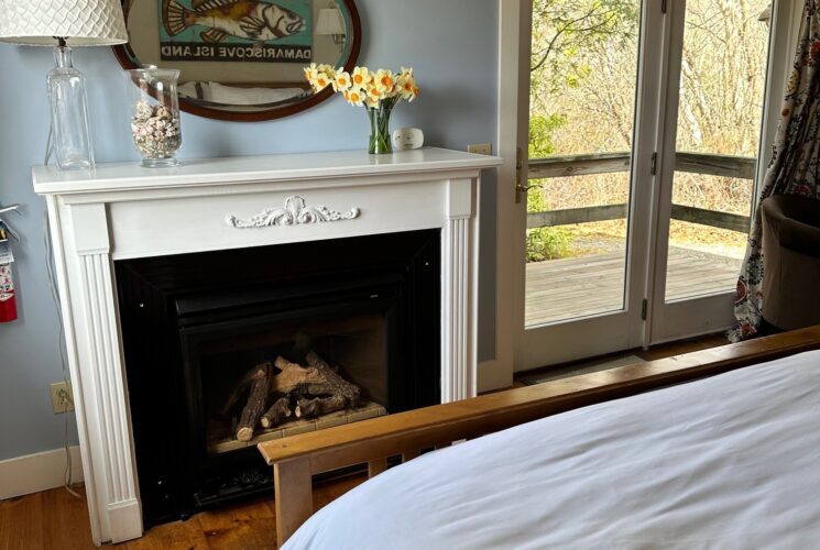 Gas fireplace with a mirror on the wall above that reflects the painting above the bed. A vase of daffodils, a glass container filled with shells and a lamp sit on the large mantle. A view of the outside deck is seen through the French door entry.