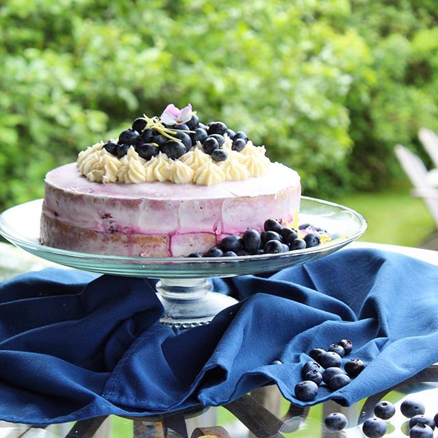 beautiful lemon blueberry cake topped with lemon buttercream and fresh blueberries on a high cake plate with greenery in the background