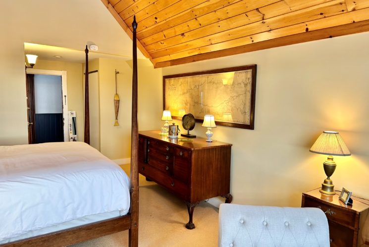 View towards the hallway with the warm lights casting a golden hue on a beautiful side board dresser topped with two small lamps and antique barometer, ice bucket and glasses. On the wall is a framed antique chart. In the view is the base of the four posted bed, part of a chair with a side table and lamp.