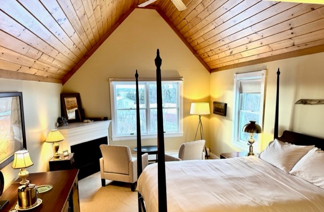 A high view of the room with its pale golden walls, high four posted bed, fireplace, chairs facing the back window looking out to the bay in the distance, warmly lit with lamps around the room