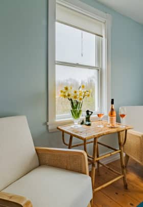 Two wood and wicker chairs with beige by the side window with daffodils and bottle of wine on table in between