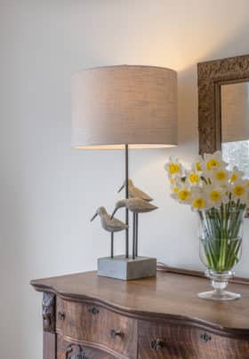 Close up of beautiful, antique sideboard dresser, sea tern lamp, daffodils, antique mirror above and vintage woman's bathing suit framed on wall.