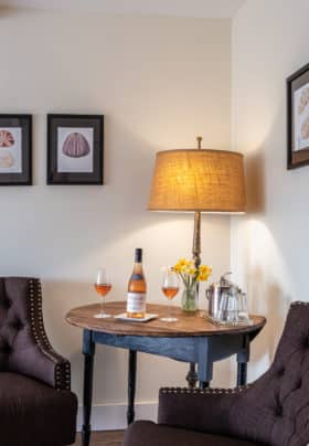 Close up view of sitting area with two chocolate brown chairs, table with standing lamp, topped with wine bottle, glasses and flowers. Five seashell prints lined up on the wall behind.
