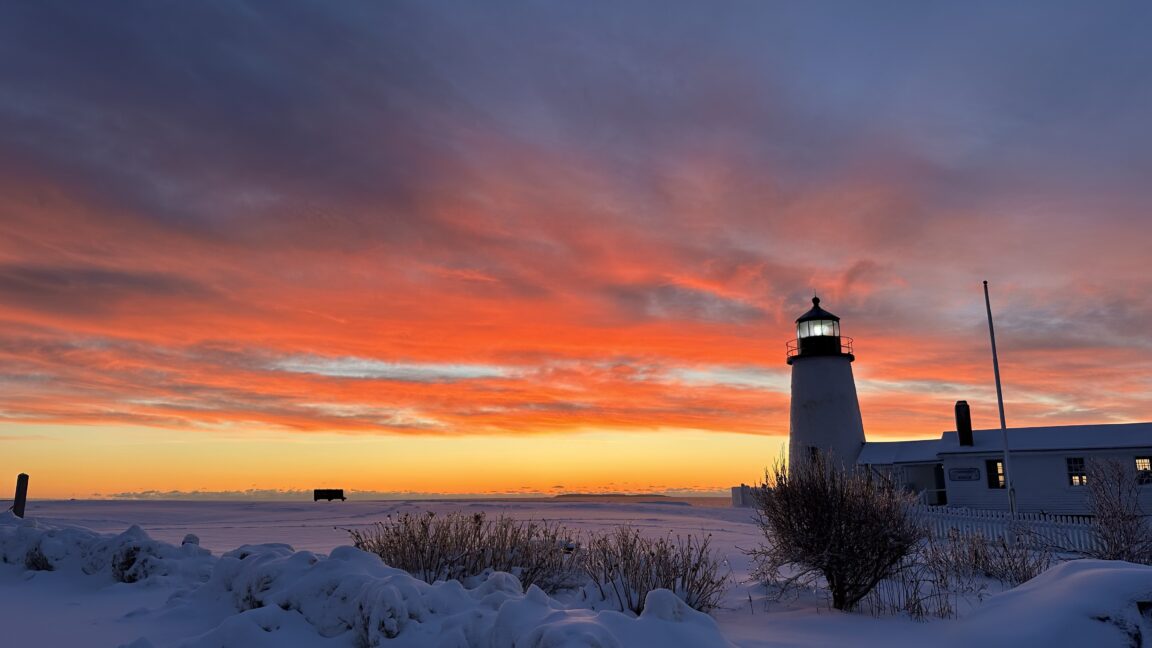 Pemaquid lighthouse at sunrise with snow on the ground. The sky is lit up with shades of orange and gray to blue clouds.
