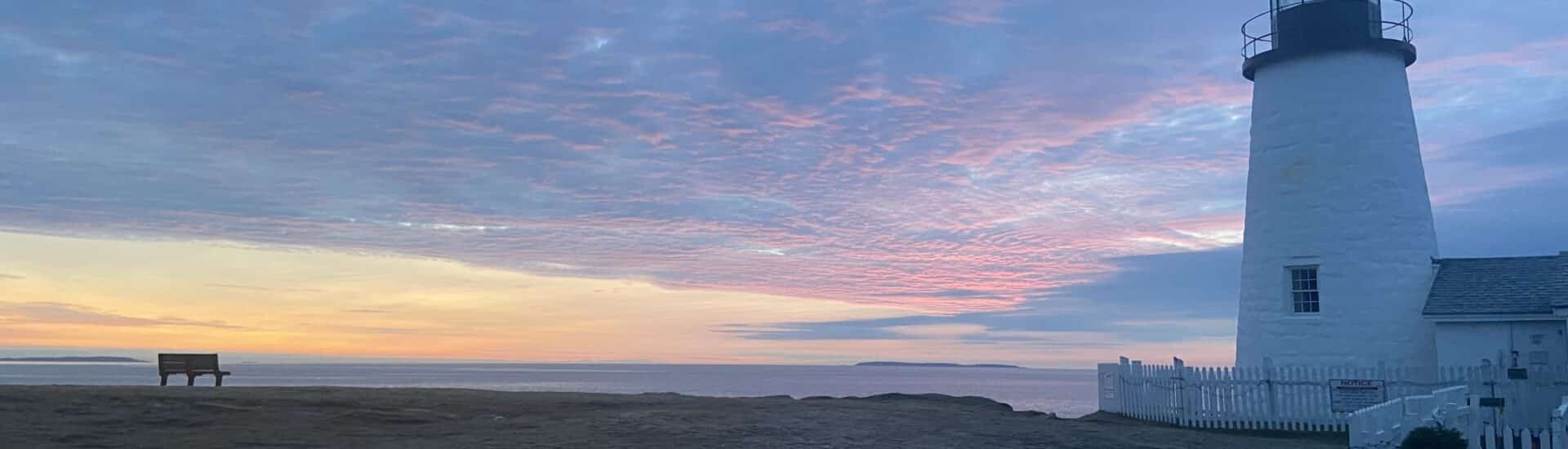 Purple clouds cover the sky with orange and yellow sunrise along the ocean. Lighthouse to the right
