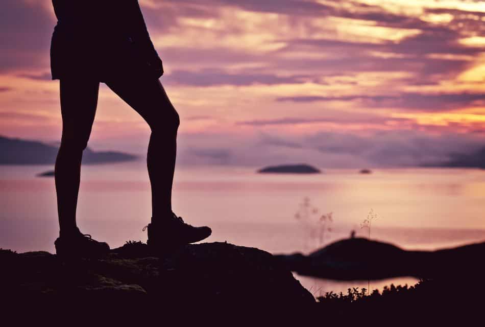 Close up view of a hiker's legs standing on a high rock with the ocean in the background at dusk