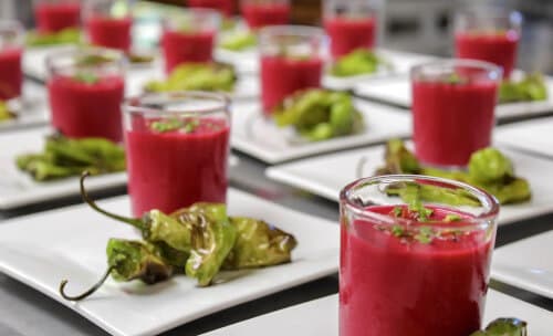 Numerous servings of beautiful hot pink beet soup in small glasses, topped with green herbs on white plates with charred shisito peppers