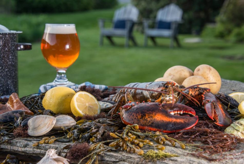 Close up view of large flat rock filled with lobster, clams, potatoes, and lemon slices and a glass of beer and large pot on the side