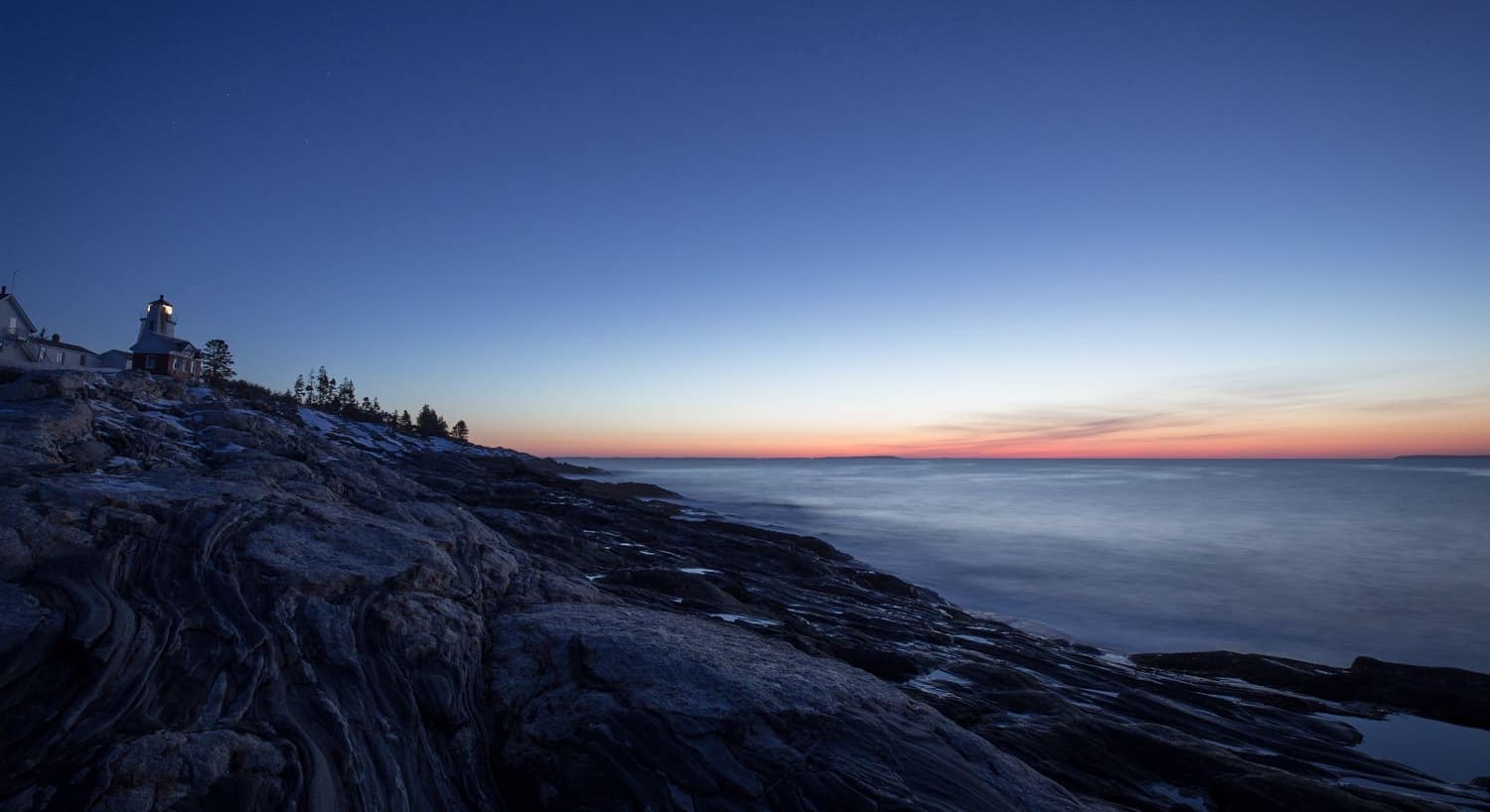 Lighthouse on a hill surrounded by large rock formations and the ocean at dusk