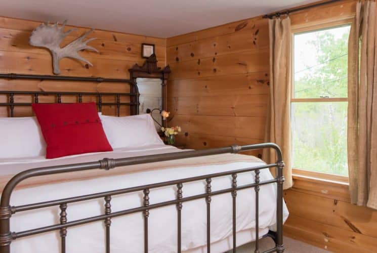 Large bedroom with wooden paneling on the walls, wrought iron bed, white bedding, and view to the outside