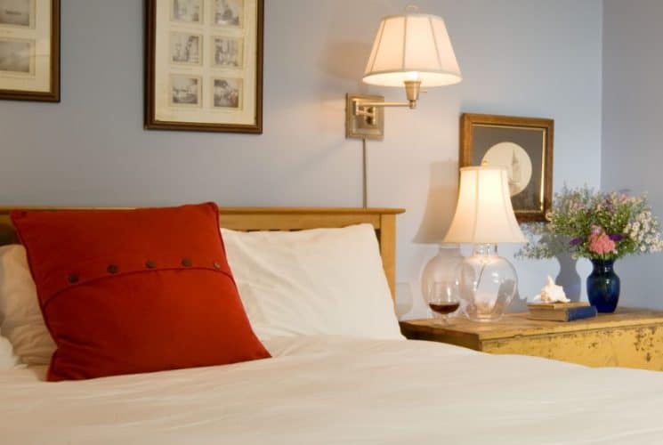 Close up view of wooden bed, white bedding, and wooden nightstand with a lamp, books, and a vase