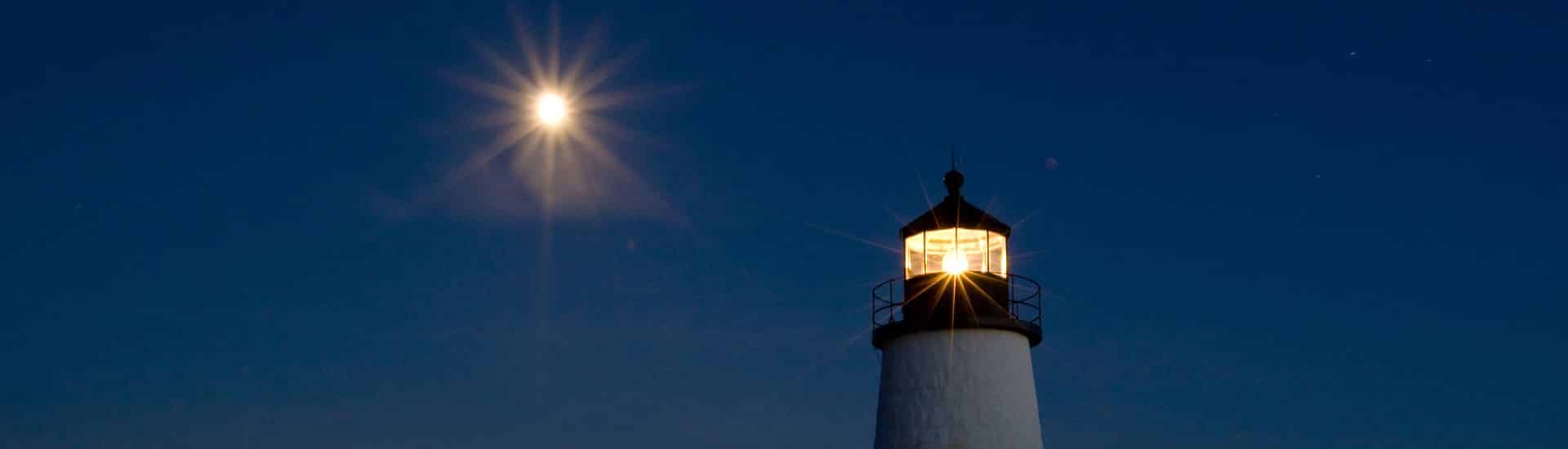 Top of a lighted lighthouse at night with moon shining in the sky
