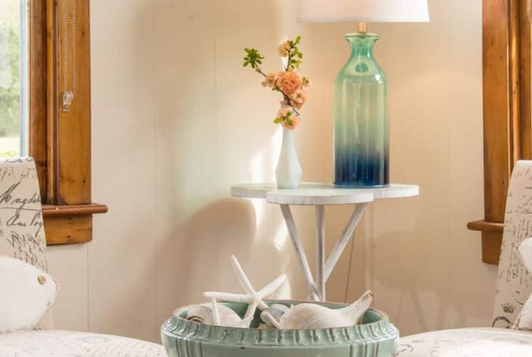 Upholstered chairs next to windows with wooden trim, small table with glass lamp and vase of flowers, and coffee table with seafoam green glass bowl with seashells