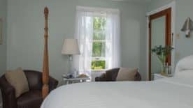 Large bedroom suite with light green walls, wooden four-poster bed, white bedding, sitting area, and view to the outside