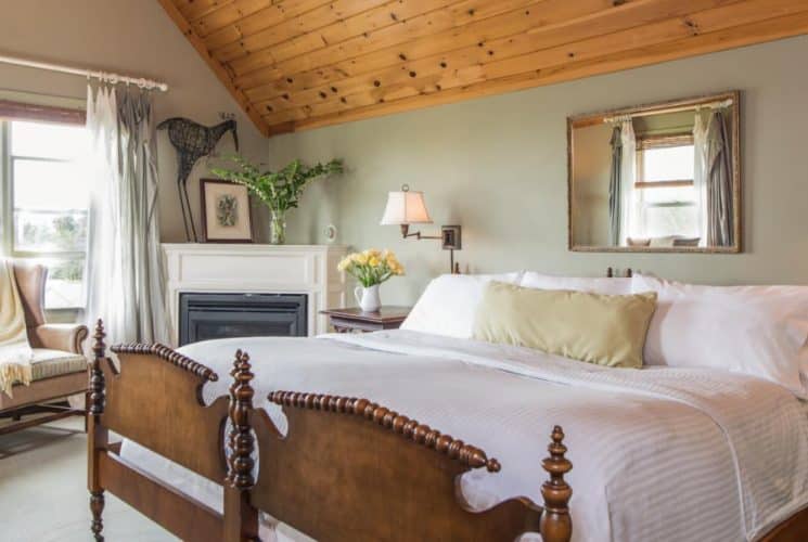 Large bedroom suite with light-colored walls, wooden paneling on the ceiling, wooden bed, white bedding, fireplace, sitting area, and view to the outside