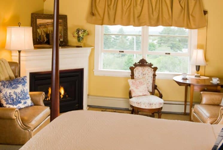 Large bedroom suite with yellow walls, wooden headboard, white bedding, sitting area, fireplace, and view to the outside