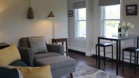 Large gathering room with light-colored walls, hardwood flooring, upholstered sofa and chair, and small table with two barstools