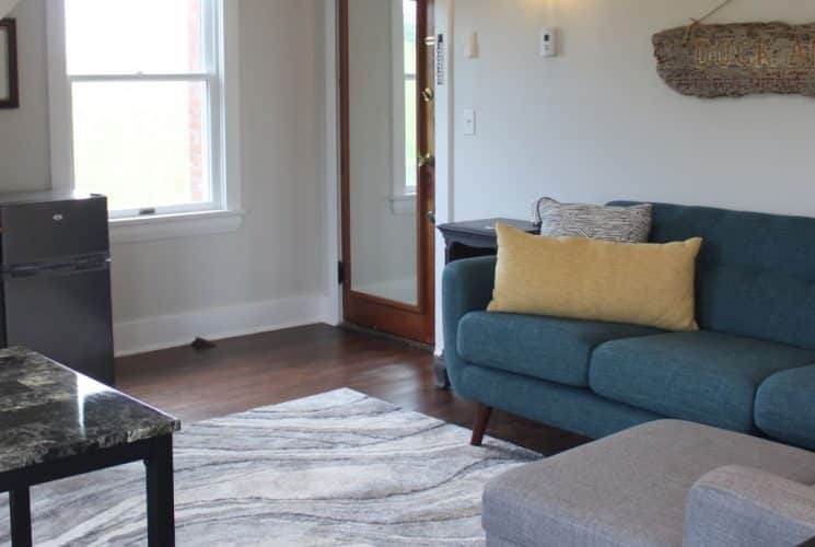 Large gathering area with light colored walls, hardwood flooring, upholstered sofa and chair, dining area, and mini fridge