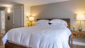 Large bedroom suite with white walls, hardwood flooring, wooden bed, white bedding, and view into the bathroom