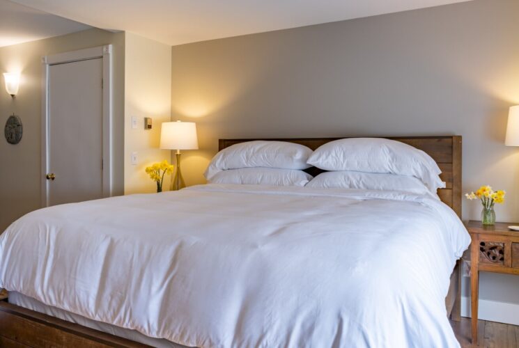 Large bedroom suite with white walls, hardwood flooring, wooden bed, white bedding, and view into the bathroom