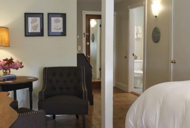 Large bedroom suite with yellow walls, hardwood flooring, white bedding, sitting area, and view into the bathroom