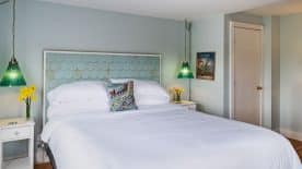 Large bedroom suite with light blue walls, hardwood flooring, white bedding, and green glass lamps hanging from the ceiling