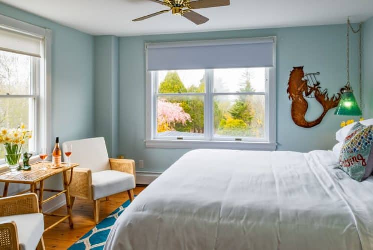 Large bedroom suite with light blue walls, hardwood flooring, white bedding, sitting area, and view to the outside