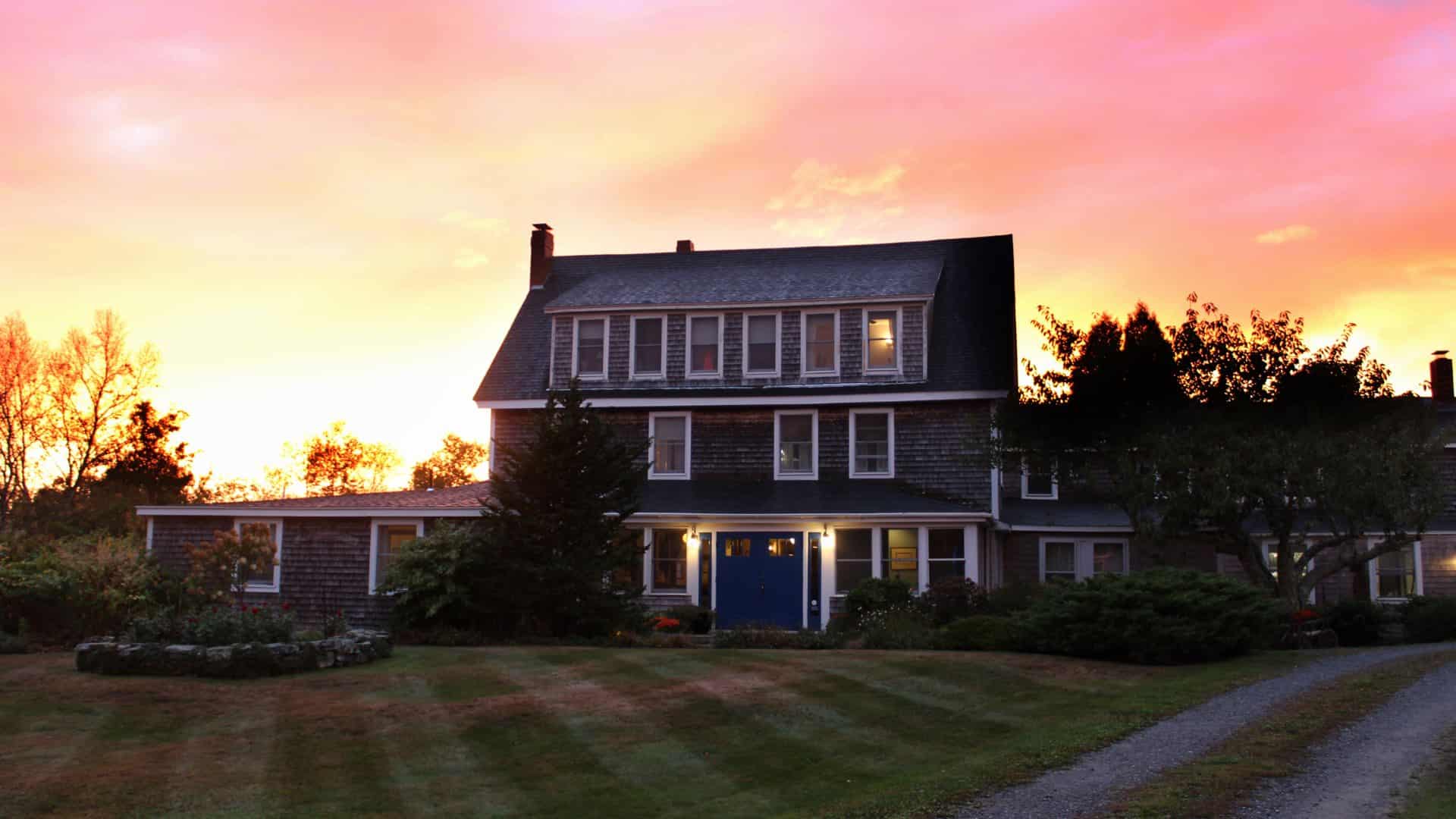 Exterior view of the property with cedar shake siding, white trim, blue door and surrounded by green grass, trees, and bushes at dusk