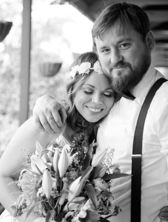 Man with white shirt and suspenders hugging woman in white dress holding a bouquet of flowers