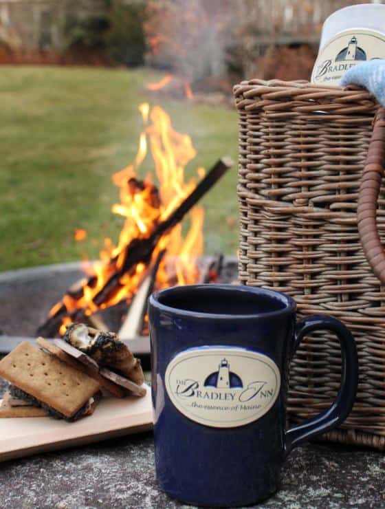 Close up view of navy stoneware coffee cup, smores, and basket with lighted firepit in the background