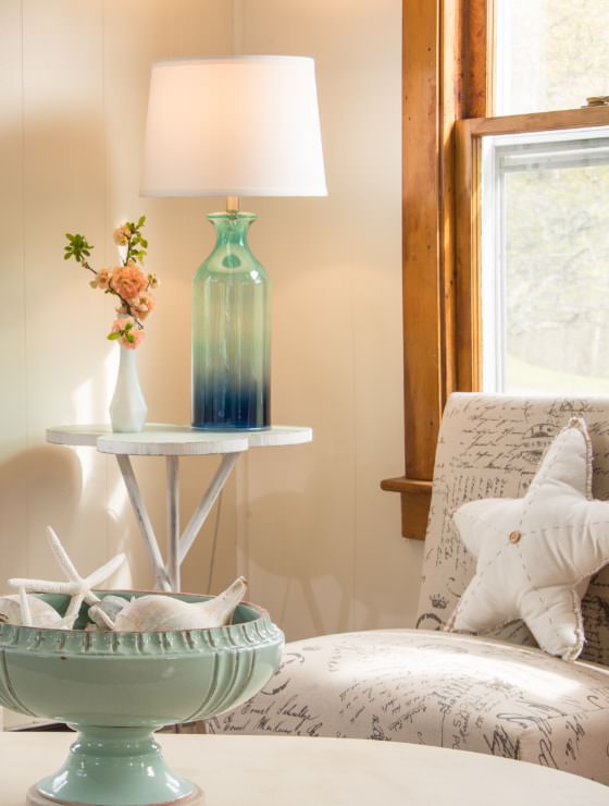 Upholstered chair next to window with wooden trim, small table with glass lamp and vase of flowers, and coffee table with seafoam green glass bowl with seashells