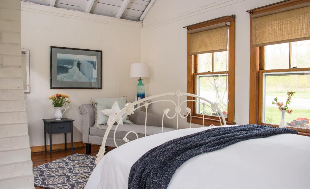 Bedroom with white walls, hardwood flooring, white wrought iron bed, white bedding, gray upholstered chaise, and windows with wooden trim