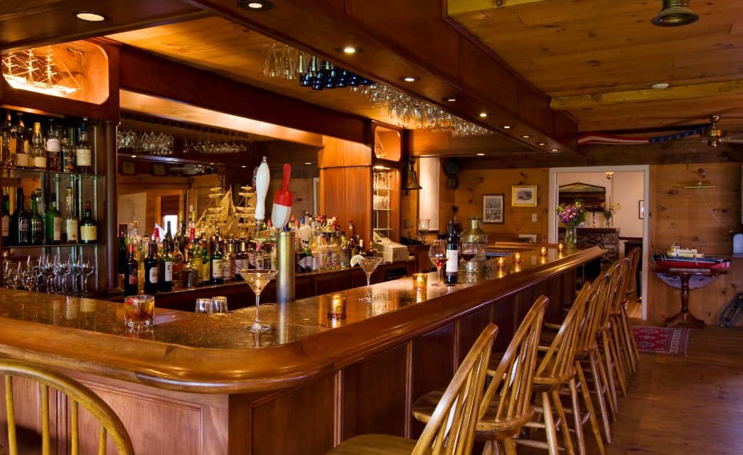 Large room with wooden paneling, hardwood flooring, wooden bar, wooden barstools, and large mirror surrounded by bottles of alcohol