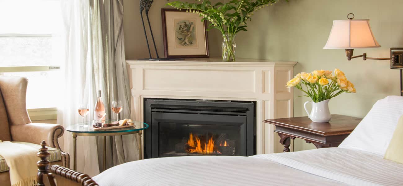 Fireplace with yellow flowers, tan wingback chair, and bed with white duvet.